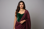 A woman wearing a dark green velvet sweetheart bustle top and a deep purple maroon pinstriped saree, styled with a gold necklace in a warmly lit indoor setting.