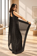 A woman models a black sleeveless maxi dress with a sheer, flowing dupatta in a warm, elegantly lit indoor setting.