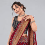A woman wearing a red and gold patterned saree with a black printed blouse, posing against a textured dark red background with gold floral motifs.