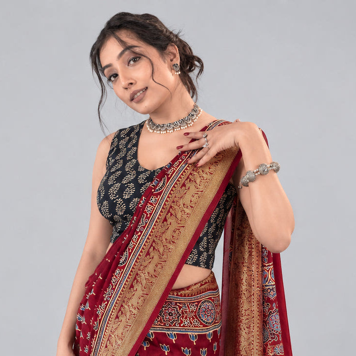 A woman wearing a red and gold patterned saree with a black printed blouse, posing against a textured dark red background with gold floral motifs.