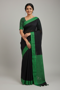 Woman wearing a black and green saree in a room with a wooden shelf and decorative items.