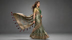 A woman modeling a flowing olive green saree with intricate gold and red patterns, a gold embroidered blouse, and a matching dupatta with red tassels, set against a dark gray studio background.