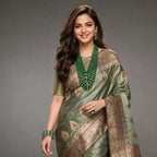 A woman modeling a sage green silk saree with intricate gold and brown zari embroidery, paired with a matching green beaded necklace, against a dark gray studio background.