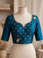 A teal blue blouse with gold embroidery, shown from both the front and back on a cream-colored dress form. The blouse features a V-neckline, elbow-length sleeves, and delicate gold motifs throughout, set against a softly blurred indoor background.